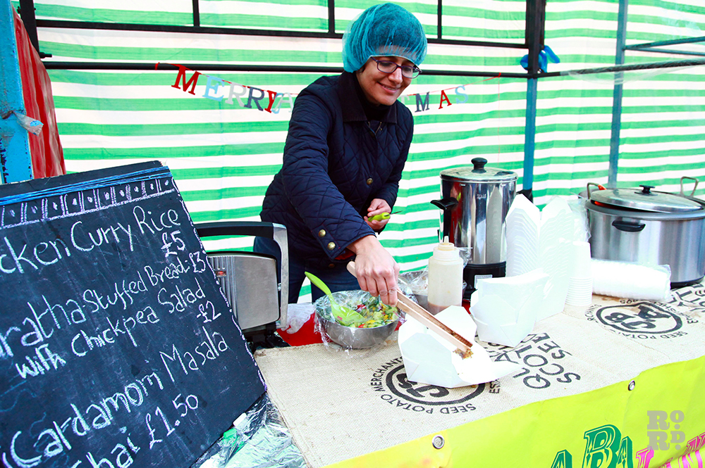 Woman in a hairnet selling Indian food at street food stall with Merry Christmas garland in felt multi-coloured lettering.