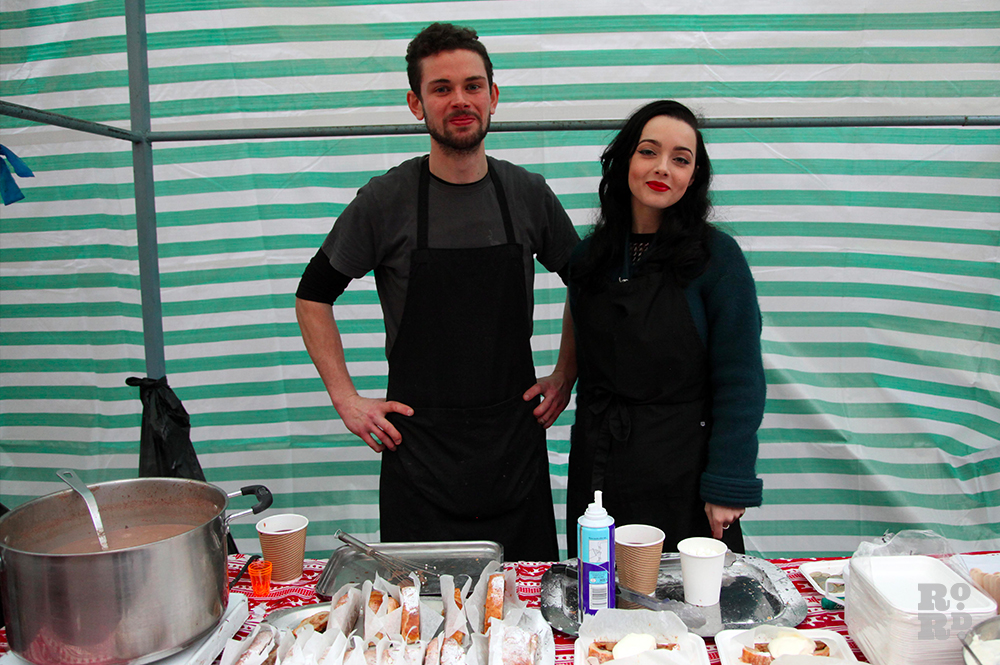 Young couple standing against green and white tarpaulin street food stall wearing black apron.