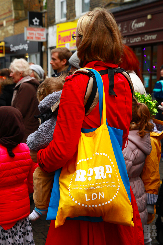 Woman in red coat with yellow and blue Roman Road LDN tote bags slung over shoulder.