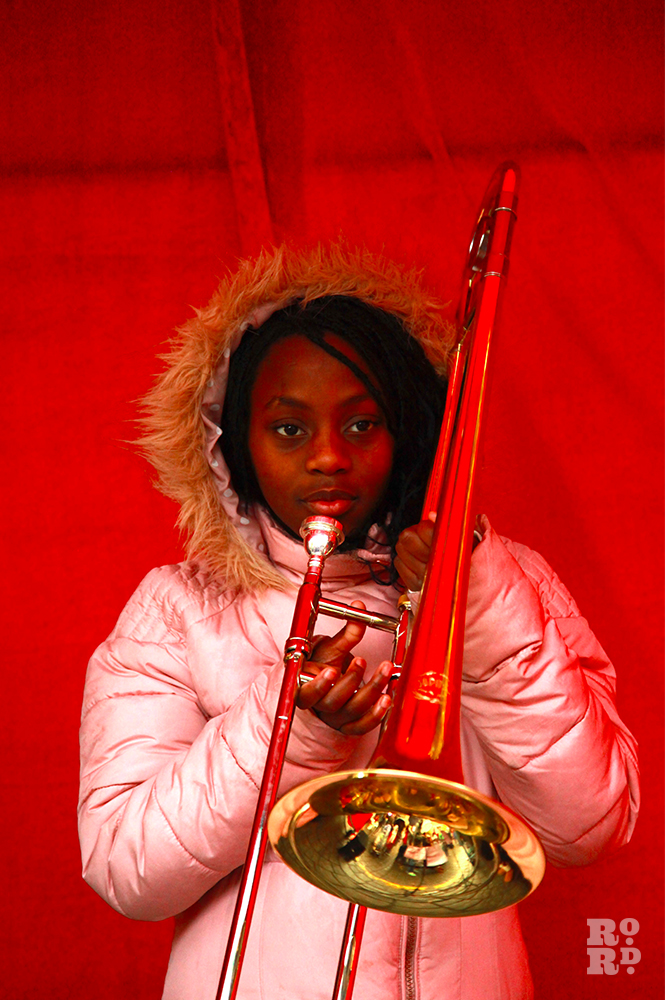 Afro-Caribbean girl in pink anorak with fur trim hood, playing trumpet against backdrop of red velvet.
