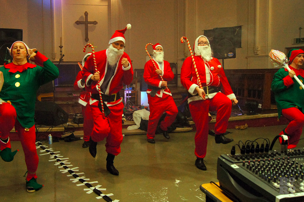Dance troupe dressed as Father Christmas holding cane, performing in a church.