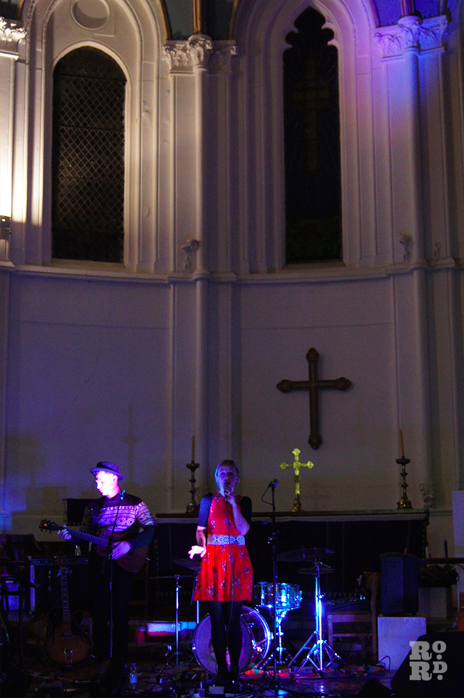 Girl in red dress singing on stage in Church, in front of large cross and tall windows.