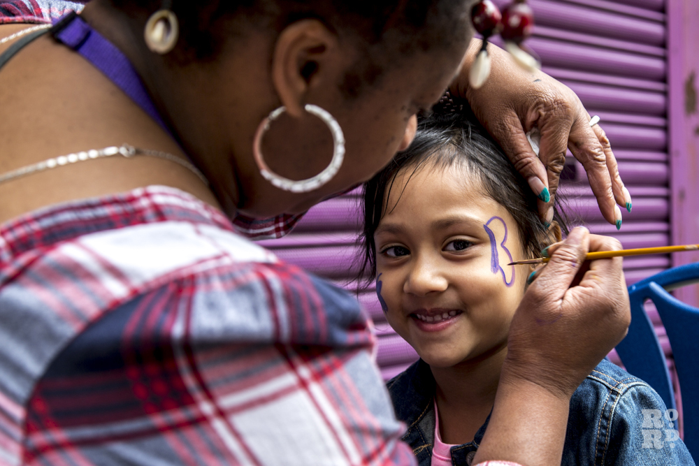 Black woman applying face paint to young asian girl at Roman Road Festival, London
