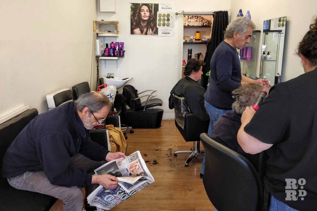 Customers getting haircuts and one waiting with newspaper at Ossie's hairdresser, Roman Road