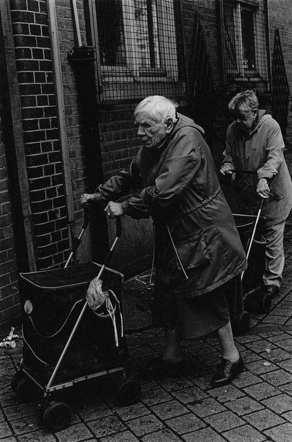 Two elderly women pushing shopping trolleys, part of a series of photographs by Stephie Devred that captures the unique spirit of the East London street market, Roman Road Market.