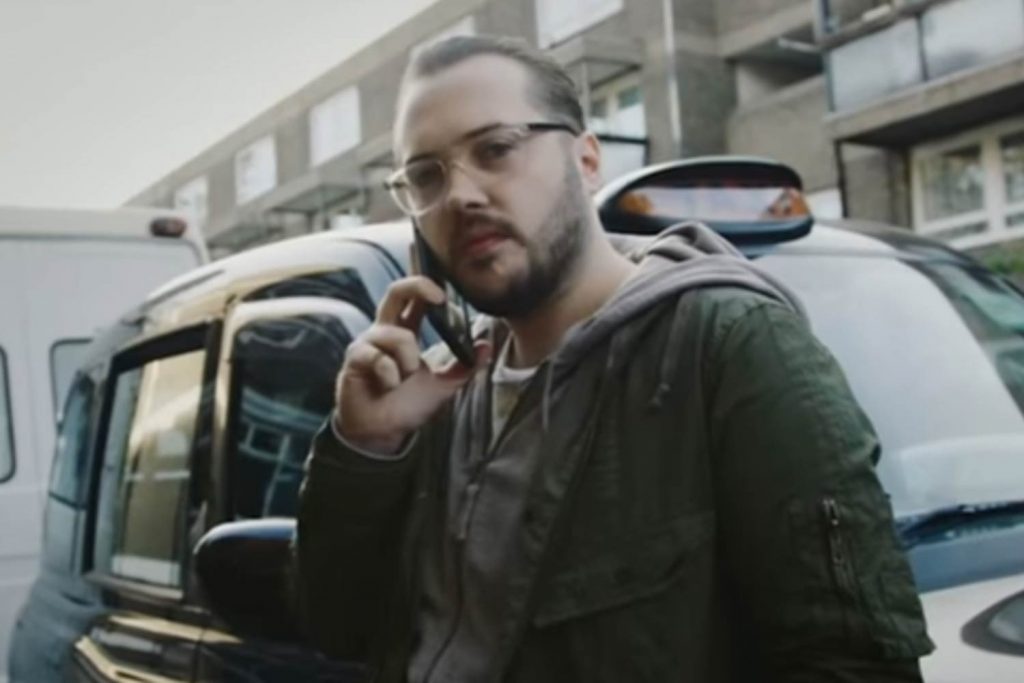 Grime videographer Risky Roadz answering a phone call in front of a London black cab near a housing estate.