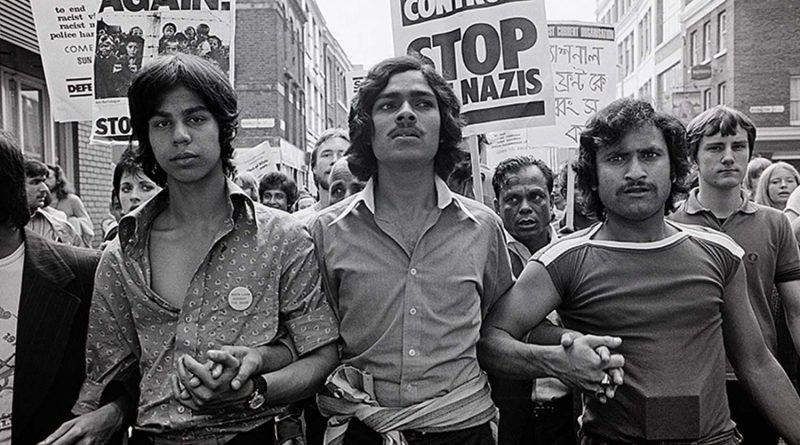 In black-and-white, three man stand arm-in-arm at the front of a protest