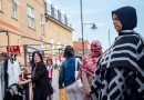 A woman with a hijab watches passers-by at Roman Road Market photoessay from 2020, by photographer Wedgley Snipes.