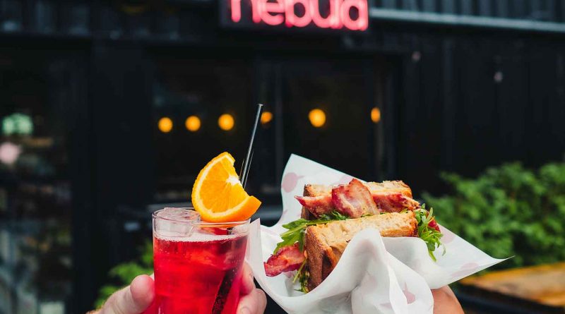 Red cocktail and bacon sandwich held in front of bar called Nebula