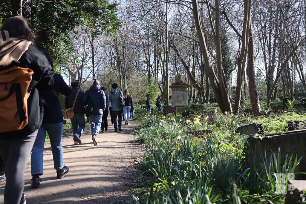 A group of people walking away from the camera, along a forested path at Tower Hamlets Bow Cemetery foraging walk