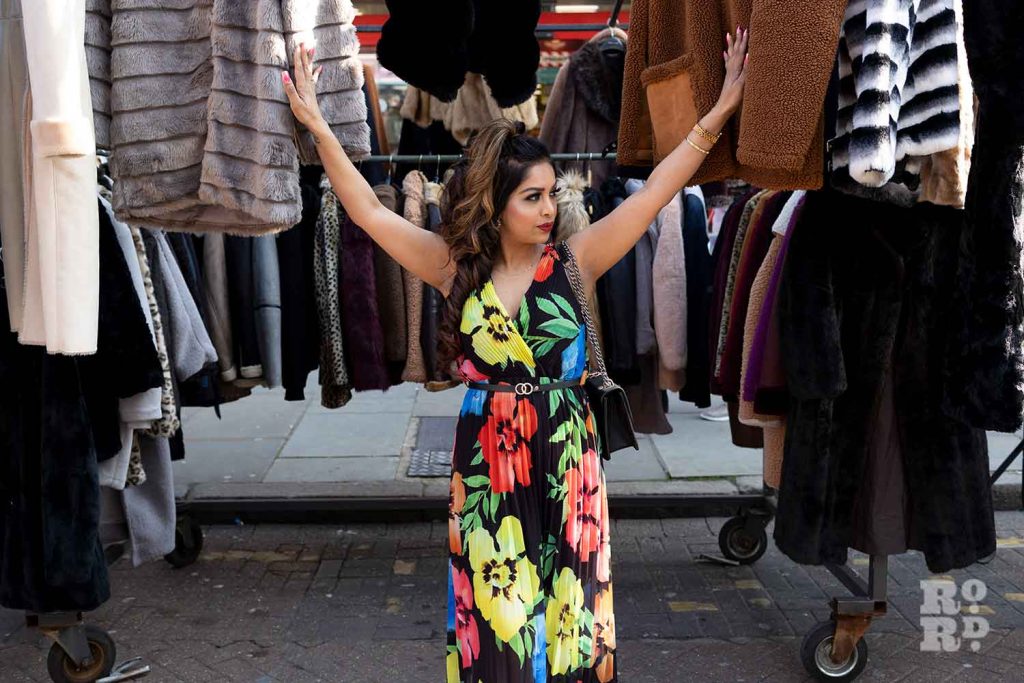 A woman in a brightly coloured floral dress posing by coats stall on Roman Road Market, Bow, East London