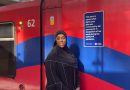 Safia Jama MBE standing in front of a DLR train with her new plaque in Stratford, East London