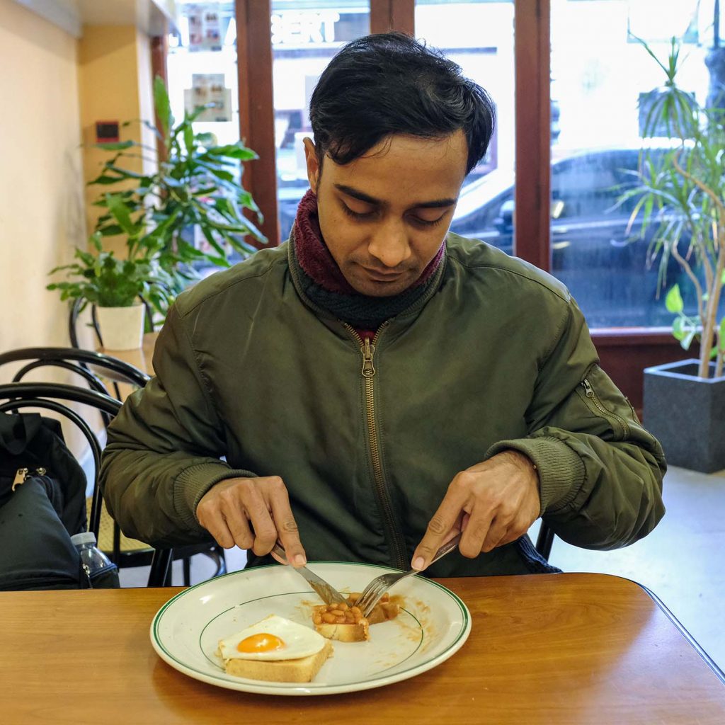 A man with scarf and coat still on eating baked beans and eggs on toast at Randolfi's, Roman Road, Bow, East London