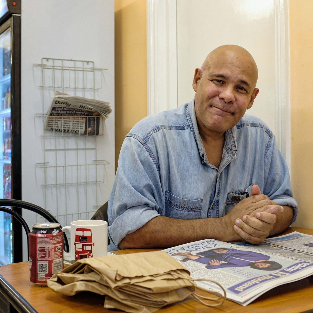 A man sitting on his own looking at the camera with a newspaper in front of him, and a can of coke, in Randolfi's, Roman Road, Bow, East London.