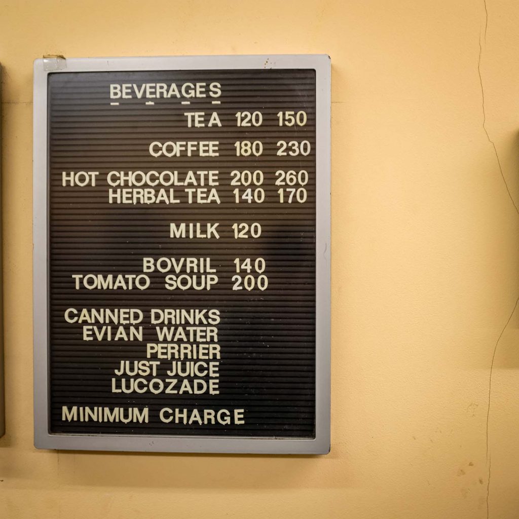 A menu of hot drinks in cream caps lock on a dark brown letterboard on a light custard yellow wall at Randolfi's, Roman Road, Bow, East London.