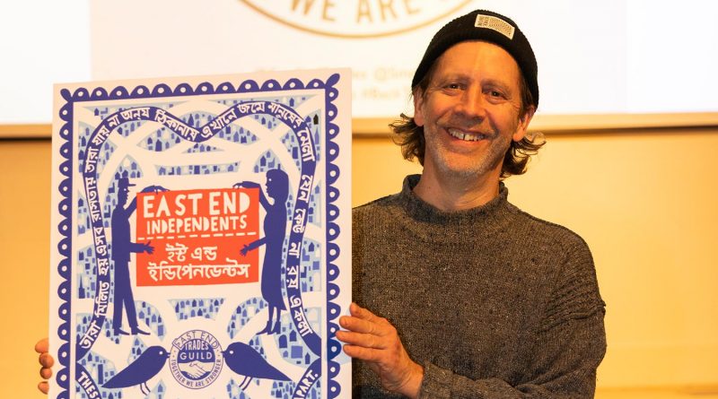 A man holding up a sign of the new East End Trades Guild guide with English writing and in Bengali, at the Bishopsgate Institute, East London