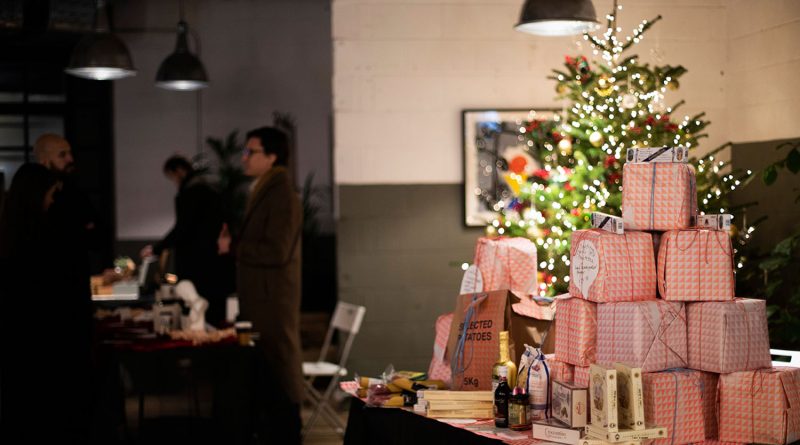 A stack of Christmas presents and a Christmas tree in the background, with people at a Christmas market stall at Redchurch Winter Market, Shoreditch, East London