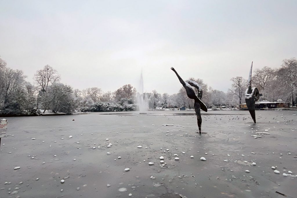 Icy lake, fountain and bird sculptures in a snowy Victoria Park, East London, December 2022.
