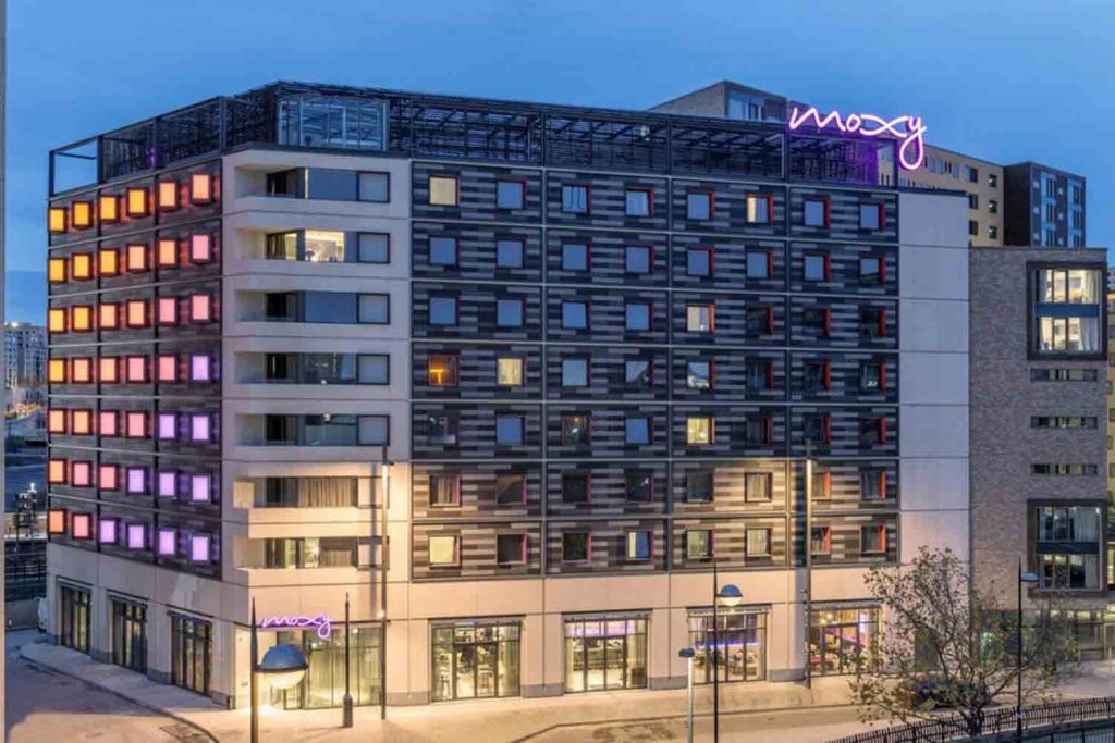 Moxy Hotel in Stratford lit up at night with neon pink sign at the building's top.