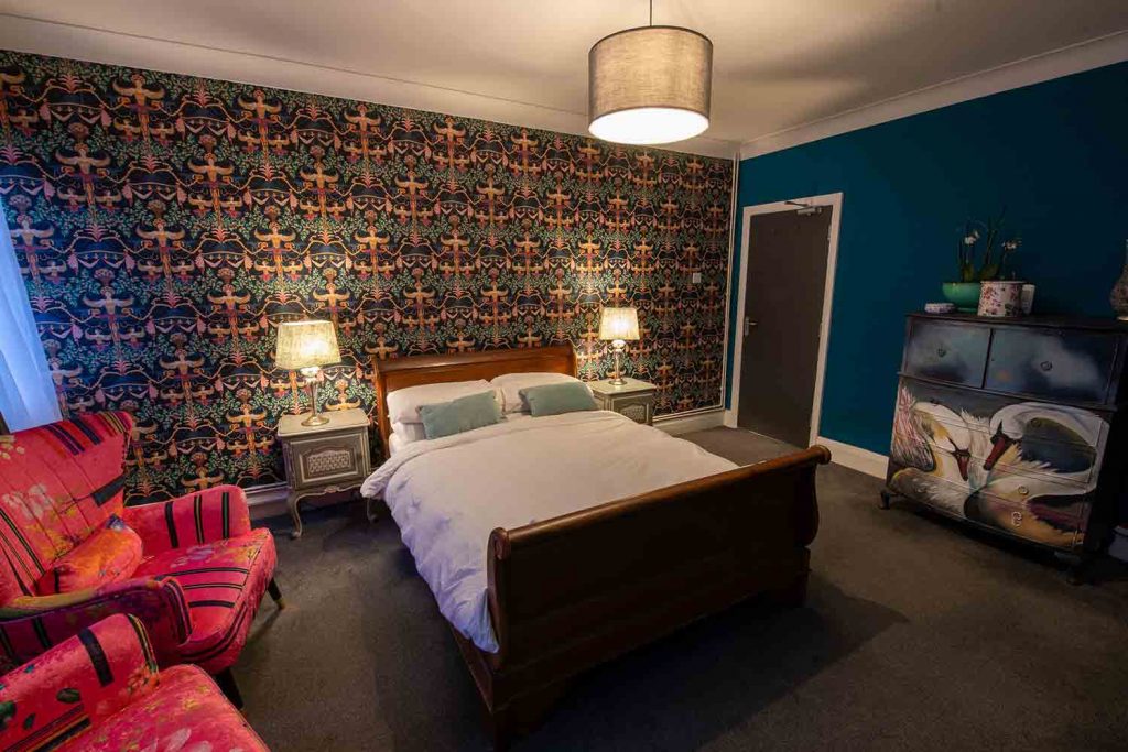 The Angel of Bow hotel inside a bedroom with dim lighting, lamps and a patterned wall decoration behind the bed.