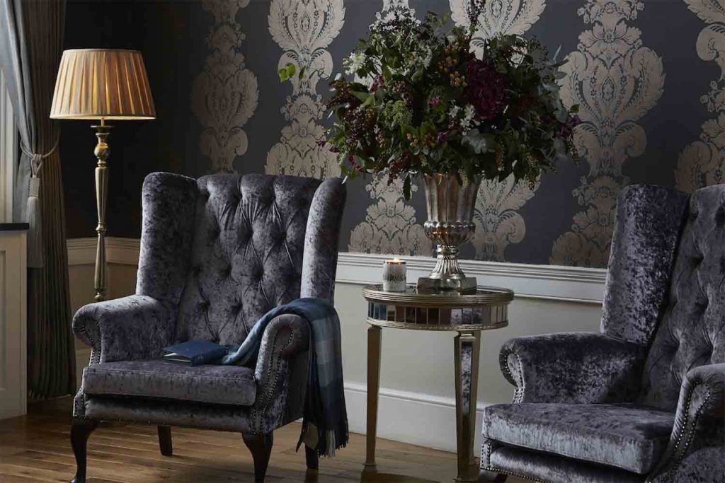 A living room inside one of the rooms of the Westbridge Hotel, with two armchairs, a yellow lamp and a house plant. Black decorative wallpaper with artistic figures.