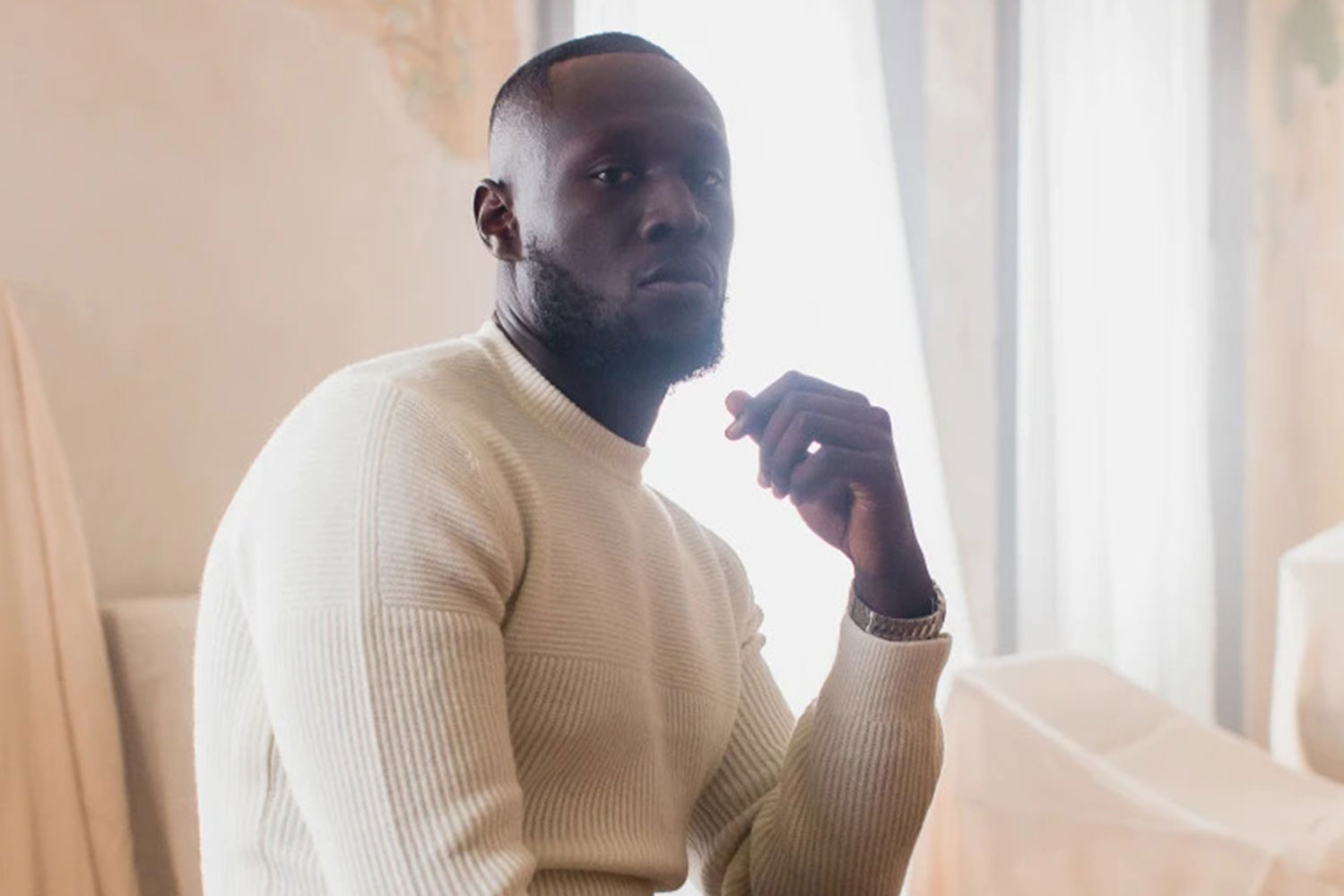Stormzy wearing a white sweater posing for a photo with an all white background.