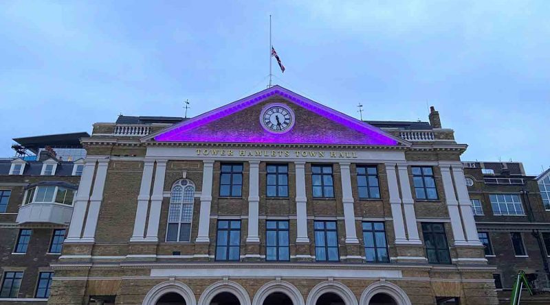 New Whitechapel town hall front with illuminated purple clock and flag above.