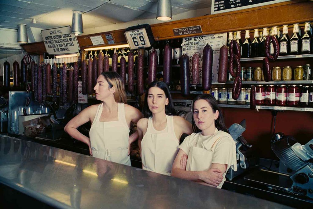 American rock band HAIM posing for a photo in front of what looks like a meat counter, with salamis and bottles in the background.