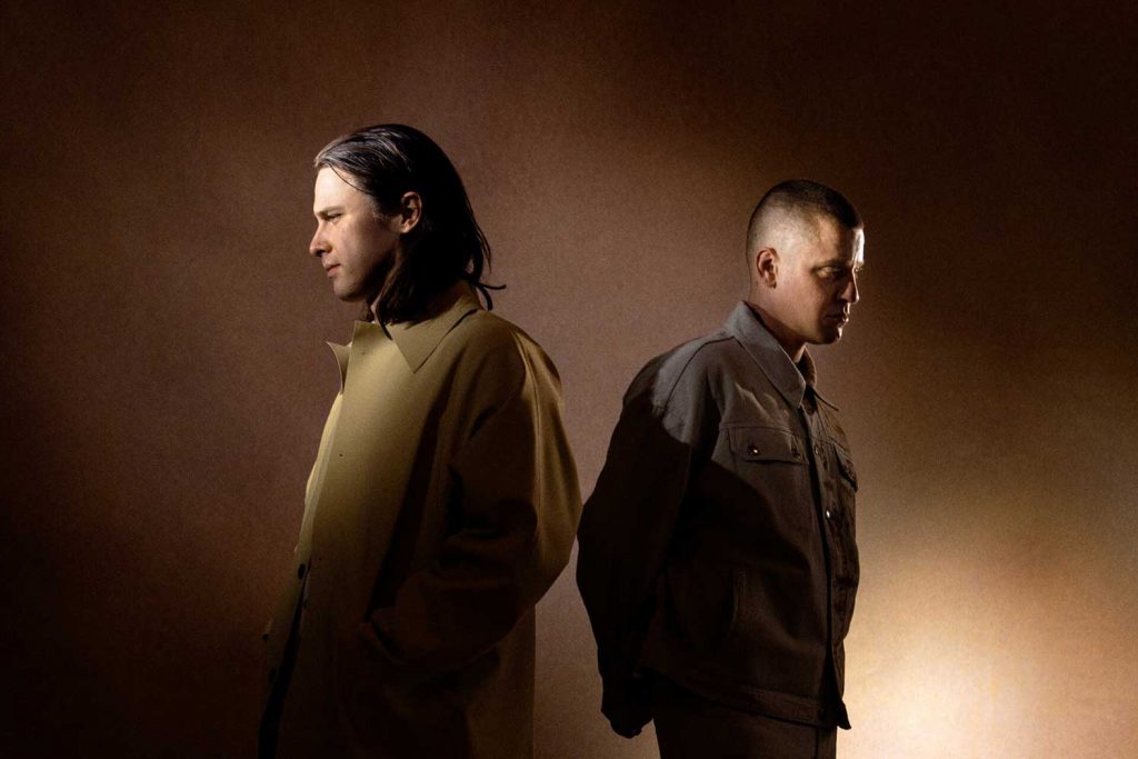 Electronic duo band Jungle posing for an artistic shot back to back against a pale black-brown background.