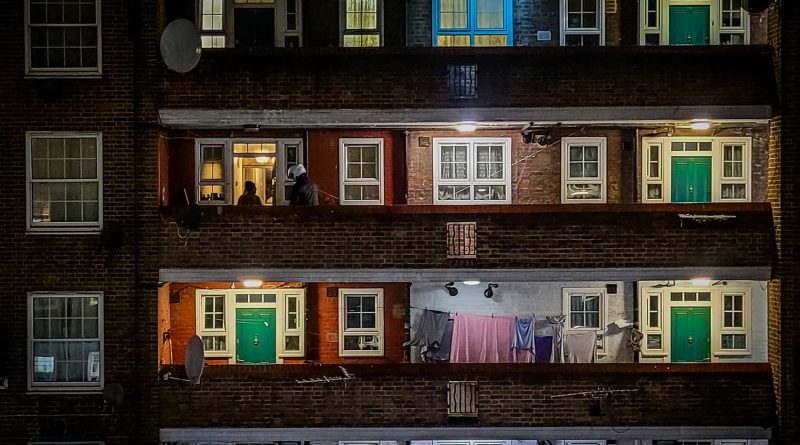 A block of flats with green doors in Whitechapel, East London, showing a courier delivering food at one of the apartments, and laundry hanging outside another. Photography by Matt Payne.