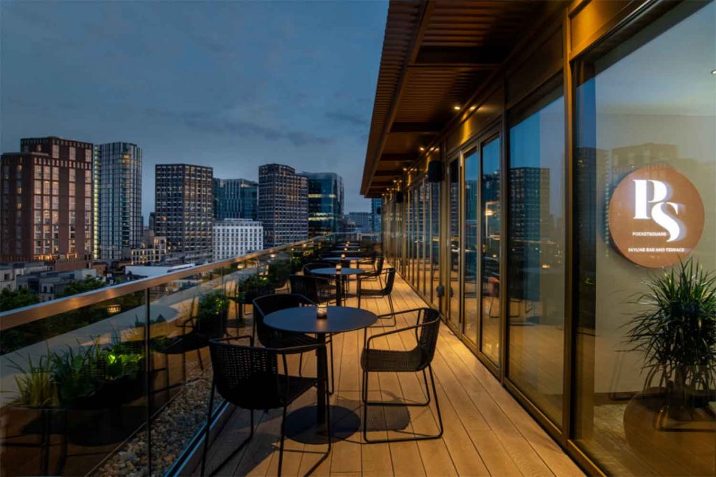 Empty rooftop bar at night with views of the city on the left and the bar through glass doors on the right.
