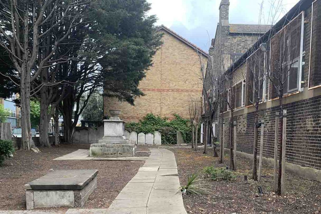 Open space surrounded by buildings with a stone path, a few trees and a few gravestones lining one of its outer perimenters