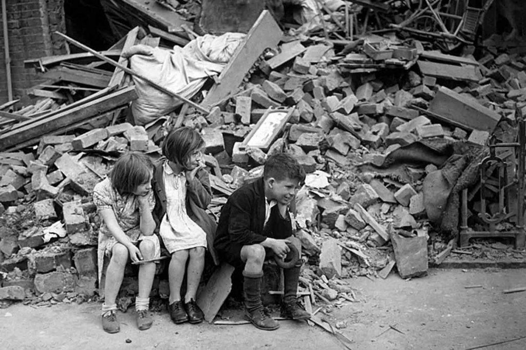 Three children sitting on rubble on a bomb damaged street in East London during World War Two.