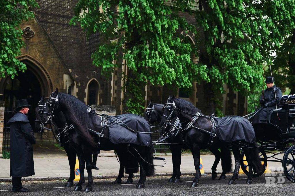Black-horse drawn funeral carriage arriving at a church in the East End.