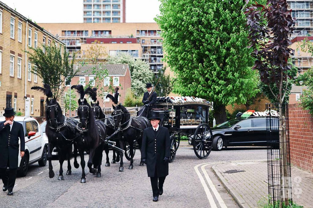 Black horse-drawn funeral carriages coming around the corner in the East End.