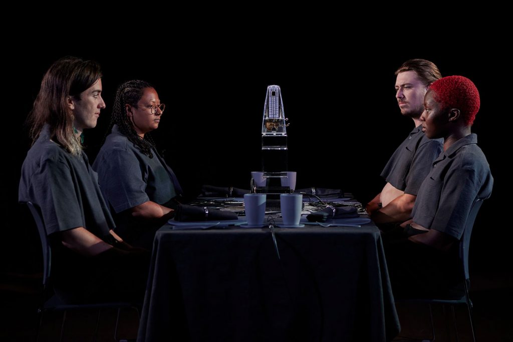 Four film actors sitting at a clinical dining table wearing matching black uniforms.