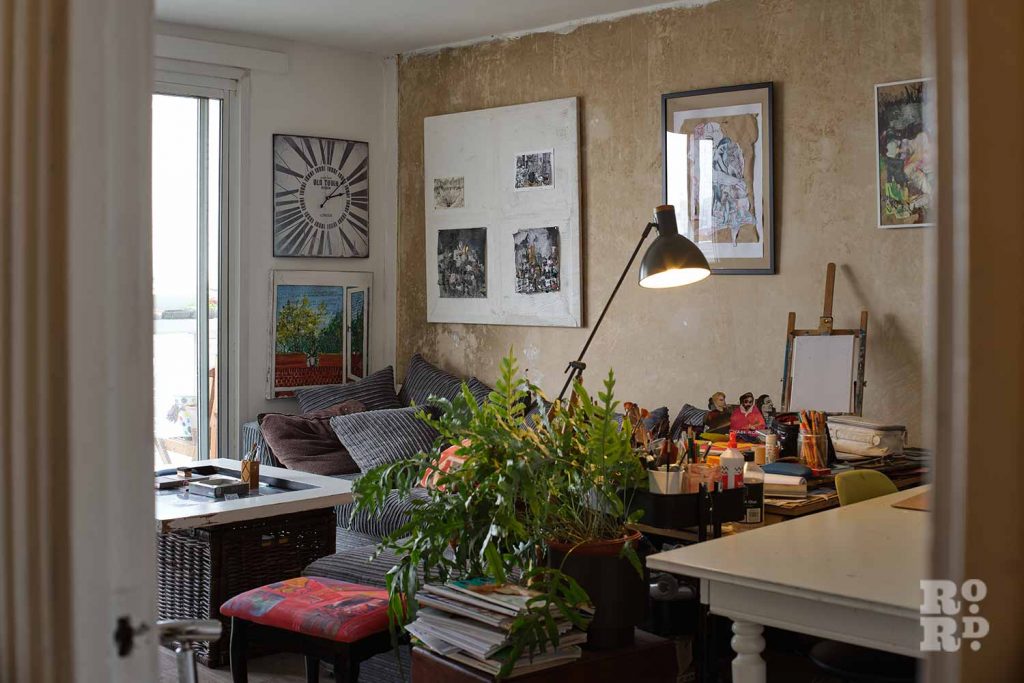 A living room with art all over the walls, paint brushes on the table, a pile of magazines and plants