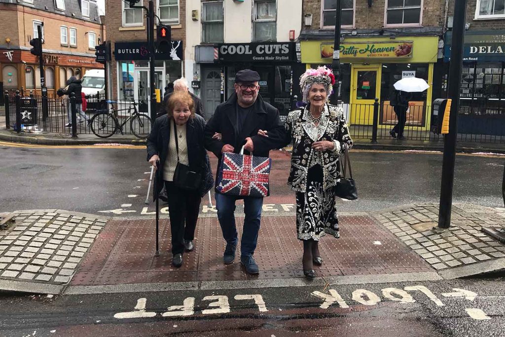 Phyllis Broadbent garbed in a mother-of-pearl outfit and pink feather hat with Geezer member Paul Dixey crossing the Roman Road