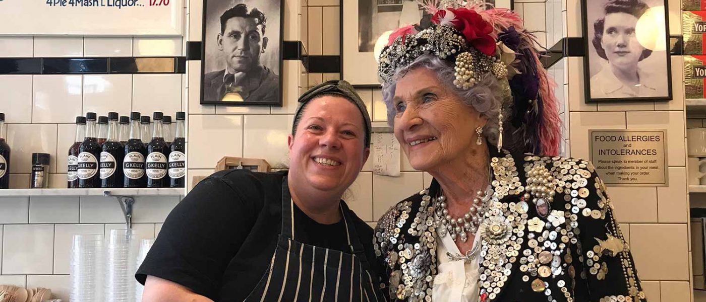 Phyllis Broadbent, the Pearly Queen of Islington, garbed in a mother-of-pearl outfit and a pink feather hat, with Leanne Black behind the counter at G Kelly's pie and mash shop on Roman Road