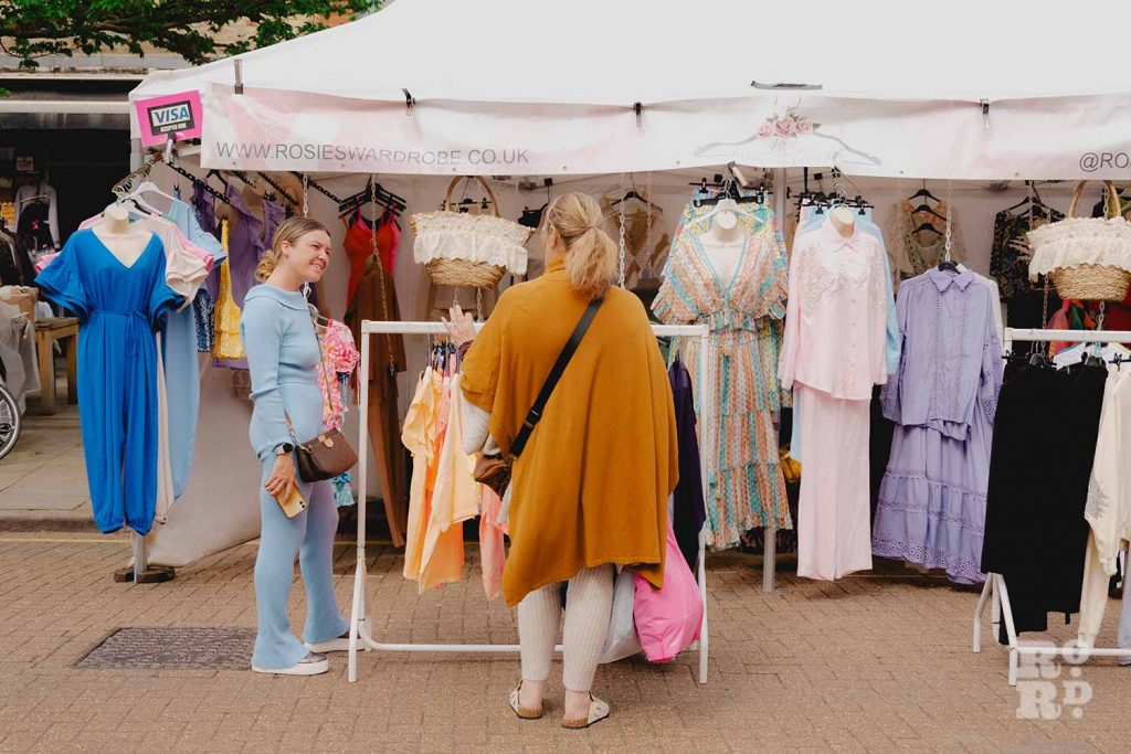Rosie Smyth wearing a blue outfit talking to a customer at her ladies' fashion stall on Roman Road Market