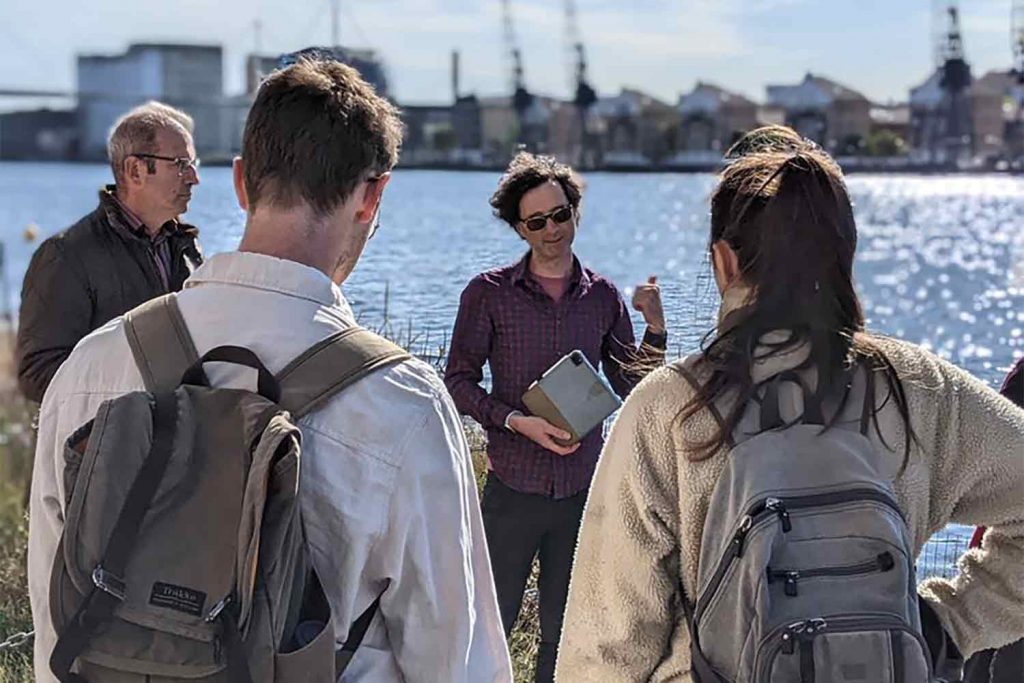 A man with an ipad describing a sunny scene on the docks to a group of interested onlookers, east London