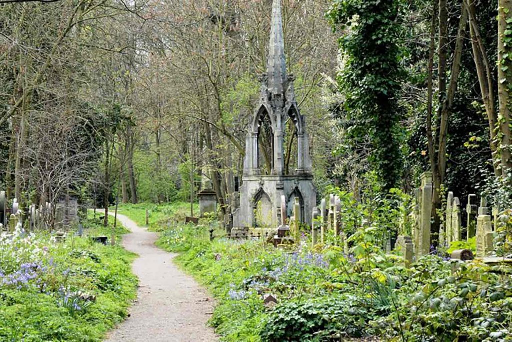 A small path leading through thick trees, flowers, and grass, surrounded by graves and a cenotaph, Tower Hamlets Cemetery Park, Bow, east London